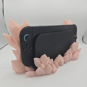May include: A black Nintendo Switch console is displayed in a light pink crystal-shaped holder. The holder has a geometric design with multiple crystal-like structures. The console is centered in the holder, with the screen facing forward.