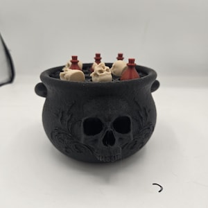 May include: A black cauldron with a skull relief on the front, filled with small beige skull figurines and red potion bottles. The cauldron has two side handles and a textured surface, holding several miniature items inside its circular opening.