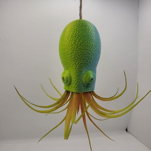 May include: A green and yellow octopus-shaped ornament hanging from a twine string. The octopus has a textured body and long, slender tentacles. The color gradient transitions from green to yellow.