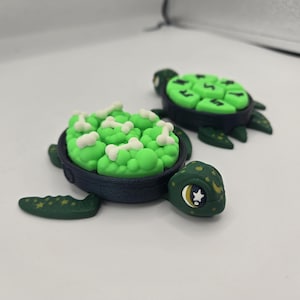 May include: Two handmade turtle figurines. One turtle has a green shell filled with white bone-shaped pieces. The other turtle has a green shell with green shapes. Both turtles have dark green bodies and heads with star and moon details.