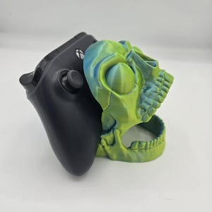 May include: A green and blue skull-shaped 3D printed stand for a video game controller.