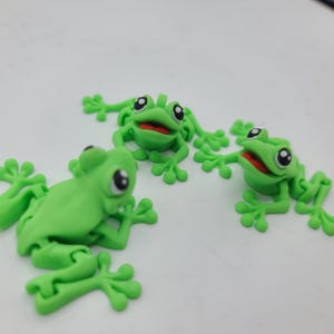 May include: Three bright green frog figurines with large black and white eyes and red mouths. The frogs are in various poses, with their limbs outstretched. The figurines are likely made of plastic or clay.