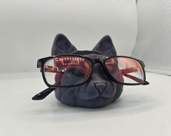 May include: A black cat-shaped glasses holder with a pair of black-framed glasses resting on it. The glasses have clear lenses with a red tint. The holder is a dark gray color and sits on a white surface.