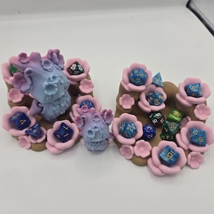 May include: Two dice holders with a fantasy theme. Each holder is brown with pink flowers and blue and purple dice. The dice are various colors, including blue, green, and black. The holders have a fantasy creature design.