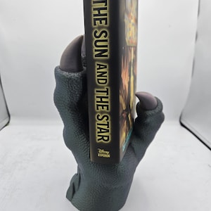 May include: A green dragon claw bookend holding a book titled "The Sun and the Star" by Rick Riordan and  illustrated by  Oshiro. The book is published by Disney Hyperion.
