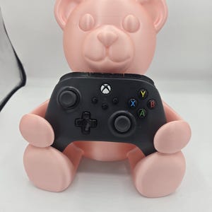 May include: A light pink 3D-printed teddy bear-shaped game controller holder. The bear is holding a black Xbox controller. The controller has colored buttons and a directional pad. The bear has a smooth, matte finish.