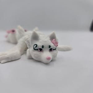 May include: A white, 3D-printed fox figurine with pink accents. The fox has a pink nose, black eyes, and a pink flower on its head. The figurine is lying down, with its paws extended.