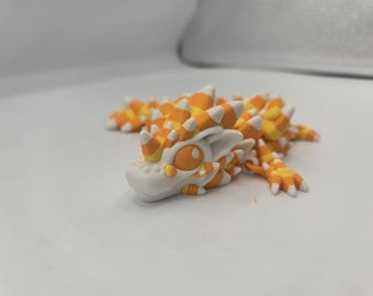 May include: A handcrafted, miniature dragon figurine in shades of orange, yellow, and white. The dragon has a detailed, textured body with spiky protrusions and large, expressive eyes. The creature is lying down on a white surface.