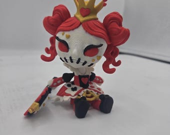 May include: A small, white, red, and gold figurine of a queen with a crown, red hair, and a red and white dress. The figurine is sitting down and holding a sword.