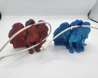 May include: Two dragon-shaped cable organizers, one red and one blue, each holding a white cable. The dragons have detailed wings and are positioned on a white surface. The red dragon has blue accents.