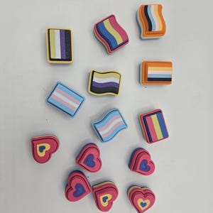 May include: A collection of colorful, small, decorative items. The items include rectangular and wavy shapes with various pride flag designs, and layered heart shapes with yellow and blue accents. The items are scattered on a white surface.