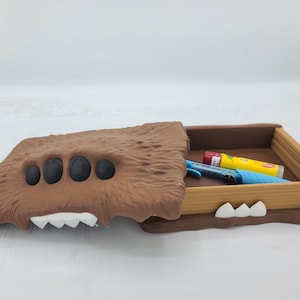 May include: A brown, monster-themed pencil box with a hinged lid. The lid has a furry texture, four black eye-like circles, and white teeth. Inside the box are several pens and lip balm tubes.