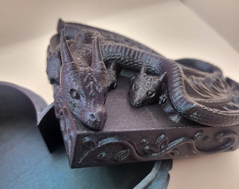 May include: A black and teal heart-shaped trinket box with a dragon and a baby dragon on the lid. The box is open and the inside is teal.