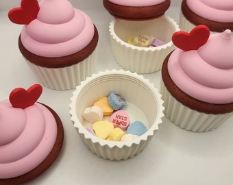 May include: A set of six white cupcake containers with pink frosting and red heart toppers. Each container has a removable lid and holds a variety of colorful candy hearts.