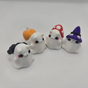May include: A collection of white ghost figurines with various Halloween-themed accessories. One ghost has a black bat-like wing, another has an orange pumpkin, a third has a red mushroom cap, and the last has a purple witch hat.