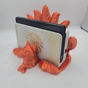 May include: Orange 3D printed stand shaped like a fox holding a Nintendo Switch console. The console is white with a gold design.