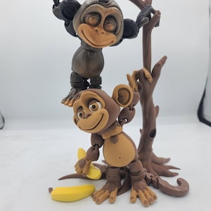May include: Two brown monkey figurines with articulated limbs, one holding a yellow banana, standing on a brown tree branch with a brown base.