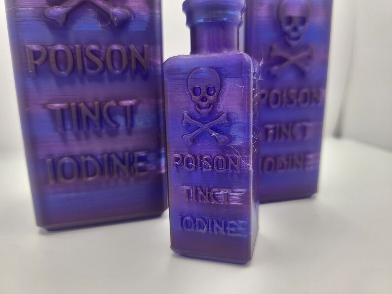 May include: Three purple glass bottles with a skull and crossbones design and the text "Poison Tinct Iodine" on each. The bottles are on a white surface.