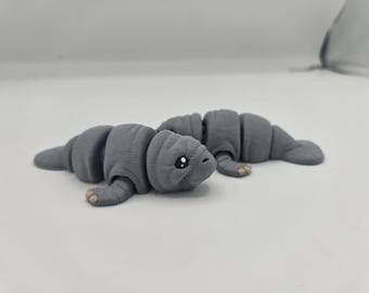 May include: A gray, segmented toy manatee with small, tan feet and a smiling face. The manatee is lying down on a white surface. The toy is made of a 3D-printed material.