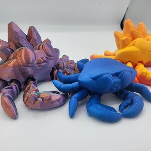 May include: Three 3D printed crab figurines. One is purple with a crystal-like design on its back, one is blue, and one is orange with a crystal-like design on its back.