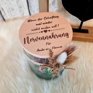 Puede incluir: Un recipiente cilíndrico de vidrio con tapa de madera. La tapa está grabada con texto en alemán y un diseño de corazón. El recipiente está adornado con flores secas y cuerda. El texto dice "Wenn der Scheißtag mal wieder nicht enden will! Nervennahrung Für Amke und Hanna."