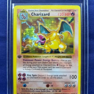 May include: A Charizard Pokémon card, Stage 2, with 120 HP. The card features a yellow and orange Charizard with blue wings, and the text "Fire Spin" with an attack power of 100. The card is in a protective sleeve.