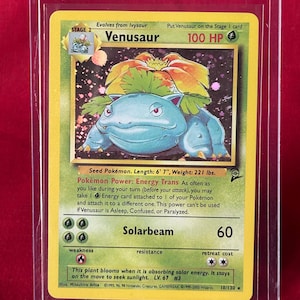 May include: A Venusaur Pokémon card in a protective sleeve. The card features a green and blue Venusaur illustration with the text "Venusaur" and "100 HP". The card's text describes its abilities and stats. The card is 6' 7" long and weighs 221 lbs.