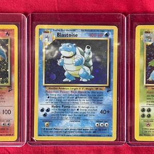 May include: Three Pokémon trading cards in protective cases. The cards feature Charizard, Blastoise, and Venusaur, each with unique artwork and game information. The Charizard card has a fire theme, while Blastoise is water-themed, and Venusaur is grass-themed.