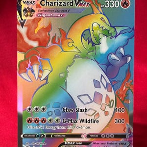 Puede incluir: Una carta Pokémon Charizard VMAX con un diseño holográfico. La carta presenta una colorida ilustración de Charizard en su forma Gigantamax, con texto que indica 330 HP y detalles de ataque. La carta se muestra sobre un soporte transparente.