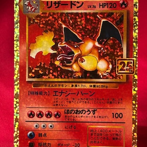 May include: A holographic Charizard Pokémon card. The card features a fiery orange dragon with large wings and a menacing expression. The card has Japanese text and a '25' emblem.