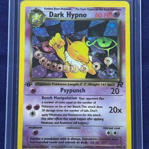 Może przedstawiać: Karta Pokémon Dark Hypno w ochronnym etui. Karta przedstawia żółte stworzenie z wahadłem na fioletowo-czarnym szachownicowym tle. Tekst karty zawiera "Psypunch" i "Bench Manipulation".
