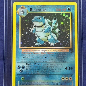 May include: A holographic Blastoise Pokémon card, Stage 2, with 100 HP. The card features a blue turtle-like creature with water cannons. The card is in a protective sleeve. The text reads "Hydro Pump Does 40 damage plus 10 more damage for each Energy attached to Blastoise."