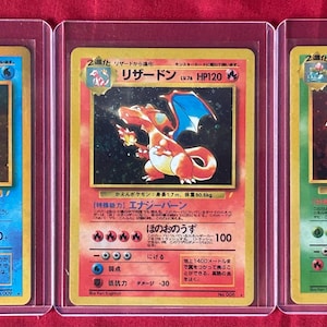 Puede incluir: Tres cartas coleccionables de Pokémon en fundas protectoras. Las cartas presentan a Blastoise, Charizard y Venusaur, cada una con texto japonés y valores de HP. La carta de Blastoise es azul, la de Charizard es naranja y la de Venusaur es verde.