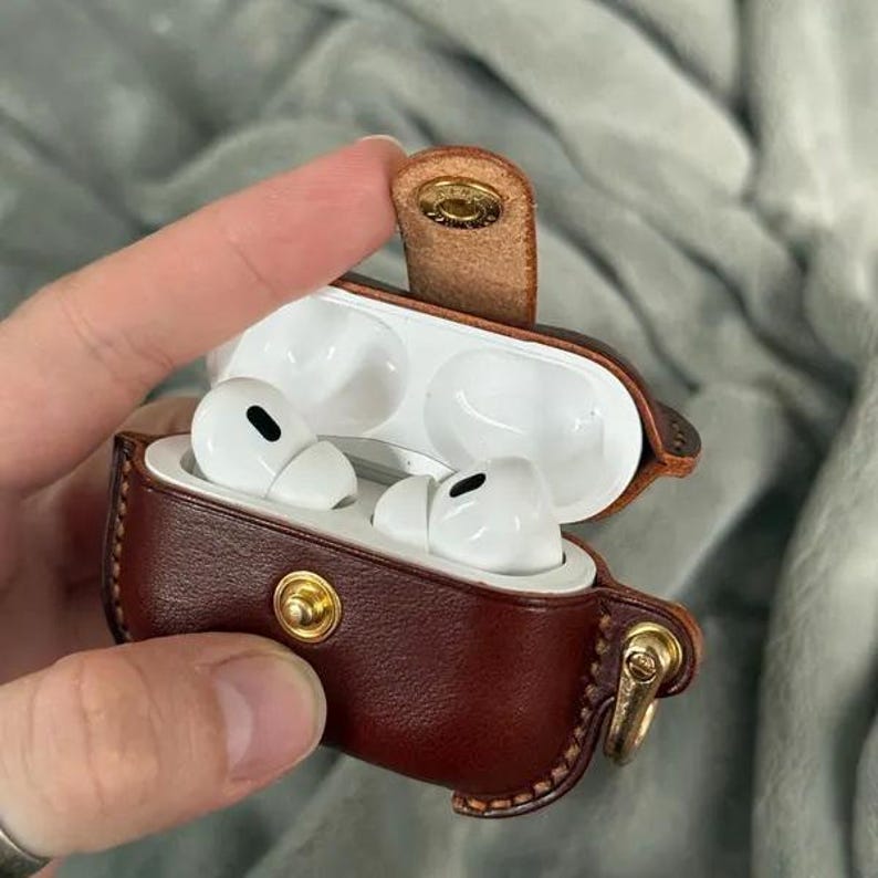 May include: A dark brown leather protective case for wireless earbuds, featuring a gold-toned metal snap button closure and a matching gold-toned side attachment ring. The case is open, revealing two white wireless earbuds inside the white plastic interior.