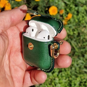 May include: A dark green leather protective case for wireless earbuds, featuring a gold-toned metal snap closure and a matching gold-toned side ring attachment. The case is open, revealing white wireless earbuds stored inside the white plastic charging unit.