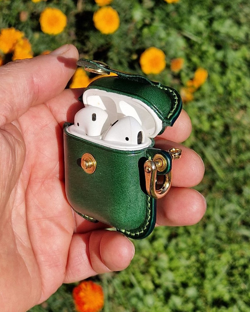May include: A dark green leather protective case for wireless earbuds, featuring a gold-toned metal snap closure and a matching gold-toned side ring attachment. The case is open, revealing white wireless earbuds stored inside the white plastic charging unit.
