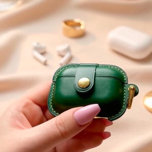 May include: A dark green protective case for wireless earbuds featuring a smooth finish, visible stitching along the edges, a central gold-toned snap button closure, and a small gold-toned metal loop attached to the side for a secure carry.