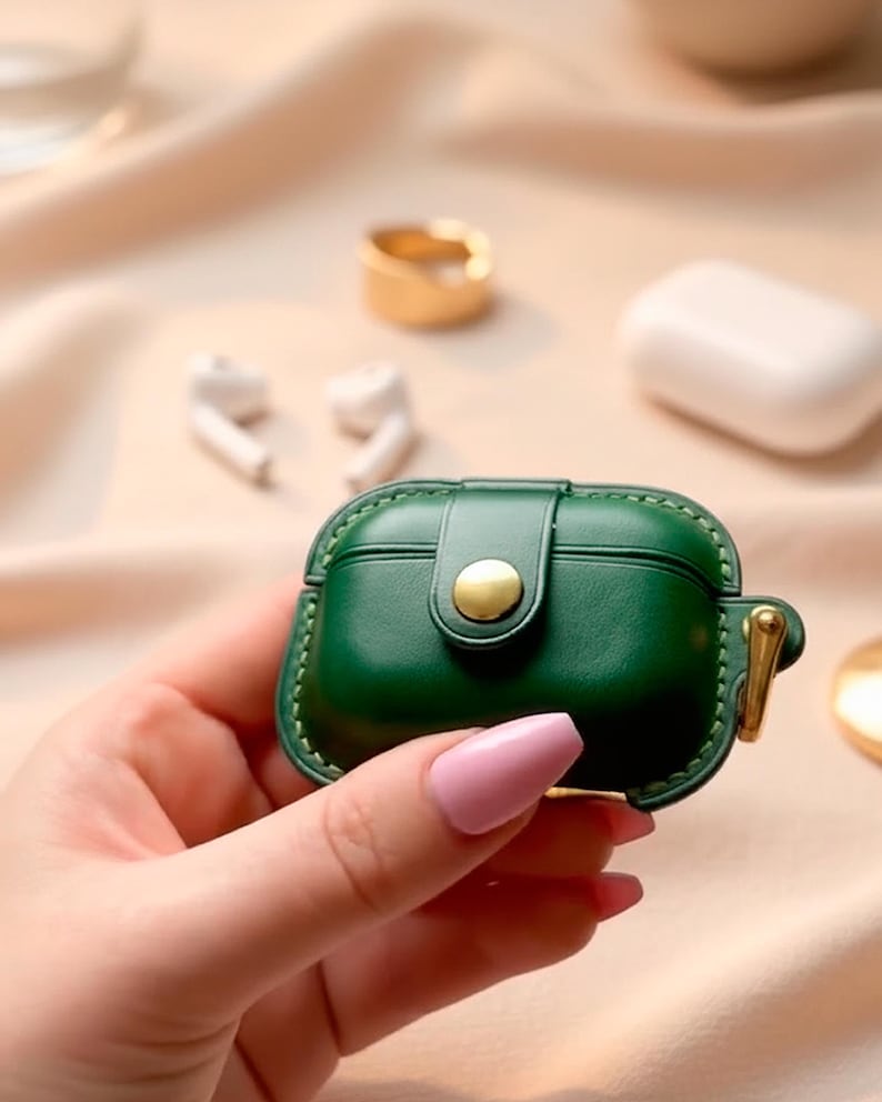 May include: A dark green protective case for wireless earbuds featuring a smooth finish, visible stitching along the edges, a central gold-toned snap button closure, and a small gold-toned metal loop attached to the side for a secure carry.