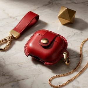 May include: A red leather protective case for wireless earbuds featuring a gold-tone snap button closure and a side attachment ring. A matching red leather wrist strap with a gold-tone lobster clasp and a thin gold-tone chain are also included.
