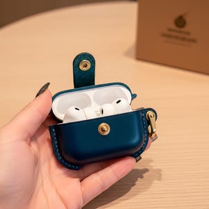 May include: A dark teal leather protective case for wireless earbuds, featuring a gold-toned metal snap button closure and a matching side attachment ring. The case is open, revealing white earbuds inside, and includes a matching tan cardboard box.