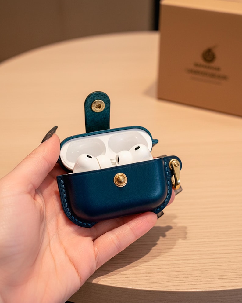 May include: A dark teal leather protective case for wireless earbuds, featuring a gold-toned metal snap button closure and a matching side attachment ring. The case is open, revealing white earbuds inside, and includes a matching tan cardboard box.