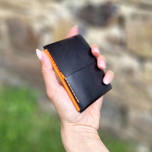 Pode incluir: Uma carteira de couro preta com costuras laranja. A carteira está dobrada ao meio e sendo segurada em uma mão.