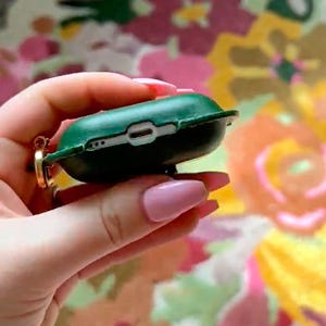 May include: A dark green protective case for wireless earbuds, featuring a cutout for a charging port and a small gold-toned metal ring attachment on the side. The case is shown from the bottom edge, revealing the charging port opening.