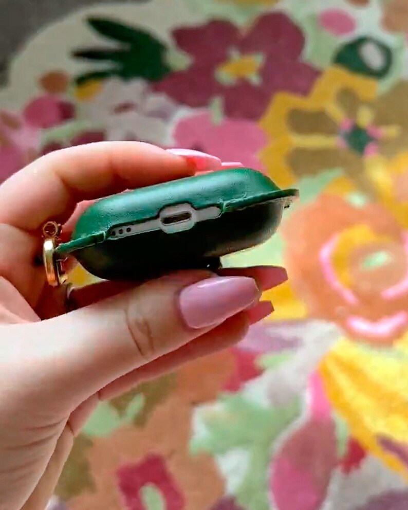 May include: A dark green protective case for wireless earbuds, featuring a cutout for a charging port and a small gold-toned metal ring attachment on the side. The case is shown from the bottom edge, revealing the charging port opening.