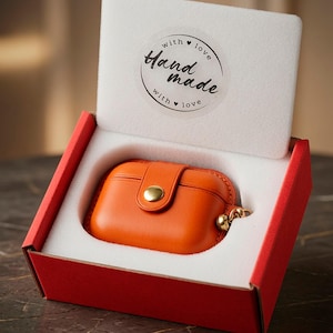 May include: An orange leather protective case for wireless earbuds featuring a gold-tone snap button closure and a small metal ring attachment, presented inside a red cardboard box with a white foam insert labelled with a Hand made with love sticker.