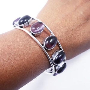 Può includere: Bracciale a polsino color argento con pietre preziose ovali, viola scuro e lavanda. Il bracciale ha un design a doppia fascia con pietre preziose incastonate in castoni individuali. Il bracciale è mostrato su un polso.