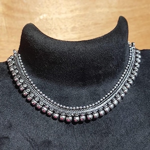 May include: A silver-toned necklace with a detailed design. The necklace features a series of small, round beads and intricate metalwork. The necklace is displayed on a black mannequin.