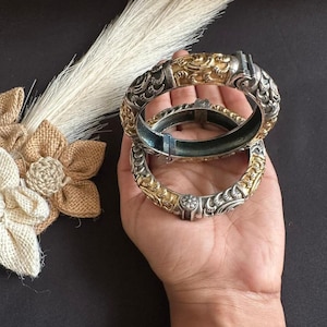 May include: Two hinged bangle bracelets featuring a two-tone design with alternating sections of polished silver-colored metal and textured gold-colored metal. The bracelets display intricate embossed patterns and a secure clasp mechanism.
