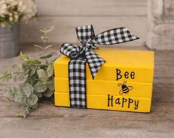May include: A stack of three yellow decorative books tied with a black and white checkered ribbon. The words "Bee Happy" and a bee graphic are printed on the front of the books. The books are on a wooden surface.