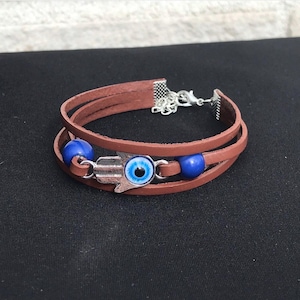 May include: Brown leather wrap bracelet with a silver Hamsa hand charm featuring a blue eye design. The bracelet also has blue beads and a silver clasp.